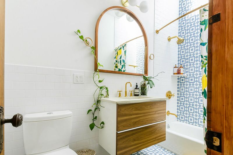 A modern blue and white bathroom with a floating wood vanity, toilet, and a shower with gold fixtures and blue and white geometric tile. There are a couple plants and an arch-shaped mirror.