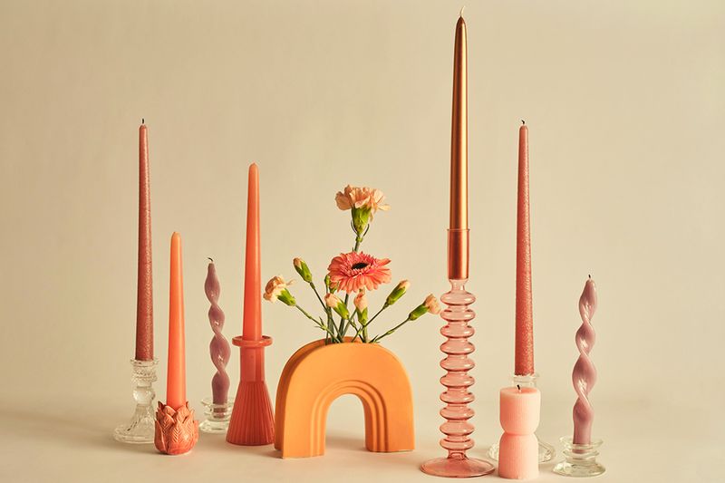 Seven taper candles of different colors and sizes (mostly orange tones) set against a beige background. All the candlestick holders are different and there's an arch-shaped flower vase in the center of the arrangement.