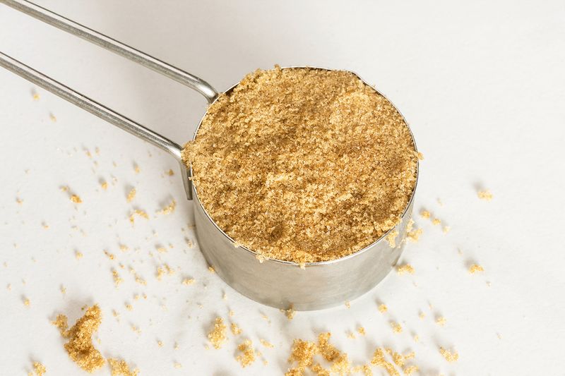 A close-up, overhead shot of a measuring cup full of brown sugar with a little bit sprinkled on the white surface it's sitting on.