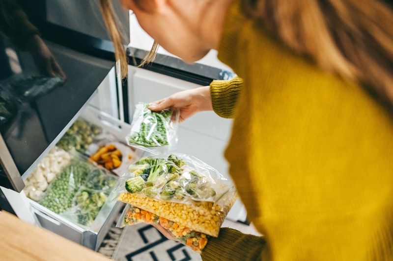 Anonymous woman wearing a mustard-colored shirt going through bags of frozen vegetables in a bottom freezer.