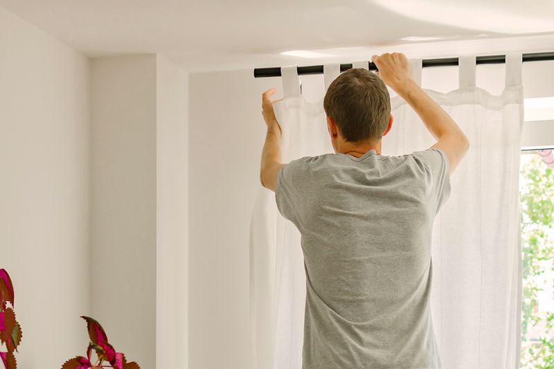 Anonymous man hanging a black metal curtain rod with white curtains on it over a window.