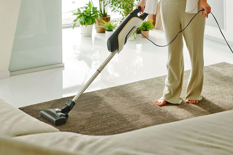 Anonymous barefoot woman vacuuming a jute runner with a diamond pattern on it. The rest of the space is white, sunny and filled with plants.