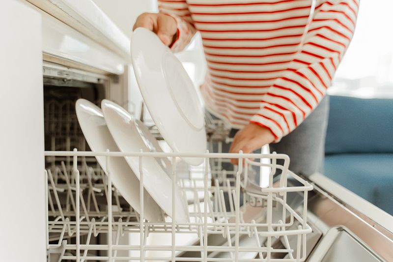 Anonymous woman in a red and white striped shirt loading plates into the top rack of a dishwasher.