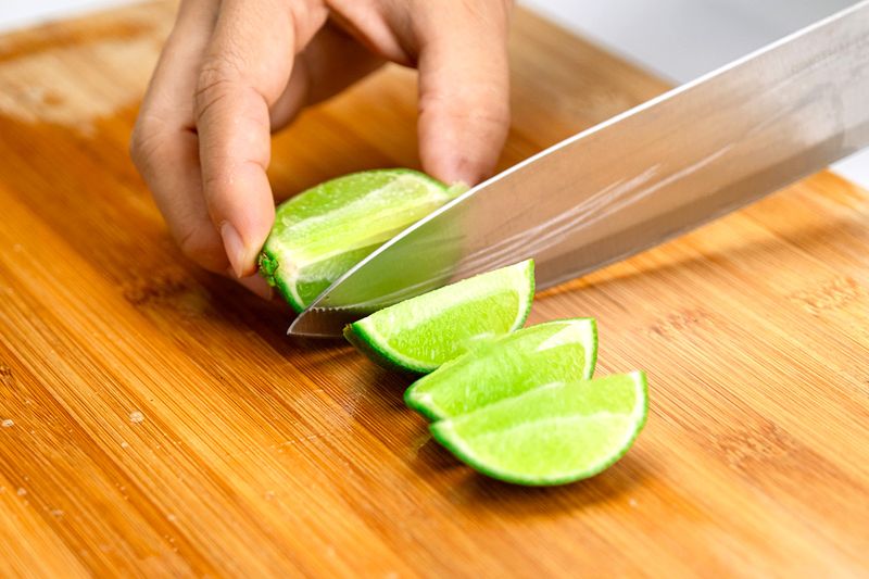 Anonymous person cutting a lime into wedges on a wooden cutting board using a chef's knife.