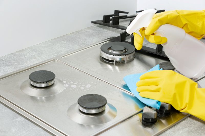 Anonymous person wearing yellow gloves while cleaning a gas, stainless steel cooktop. The person is using a cleaner in a spray bottle and a microfiber cloth to wipe down the surface.
