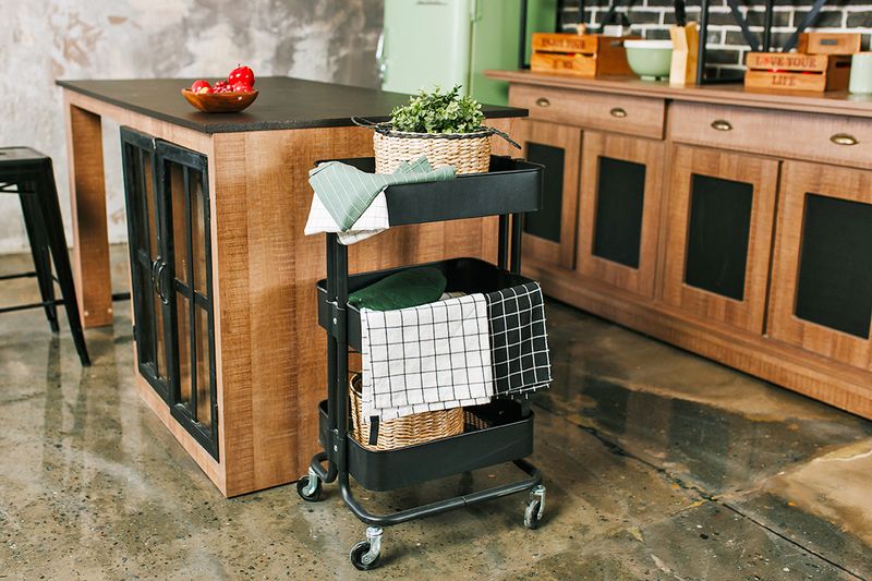 A kitchen with wood cabinets, stone floors, and a black metal three-tier rolling cart with cooking essentials inside of it.