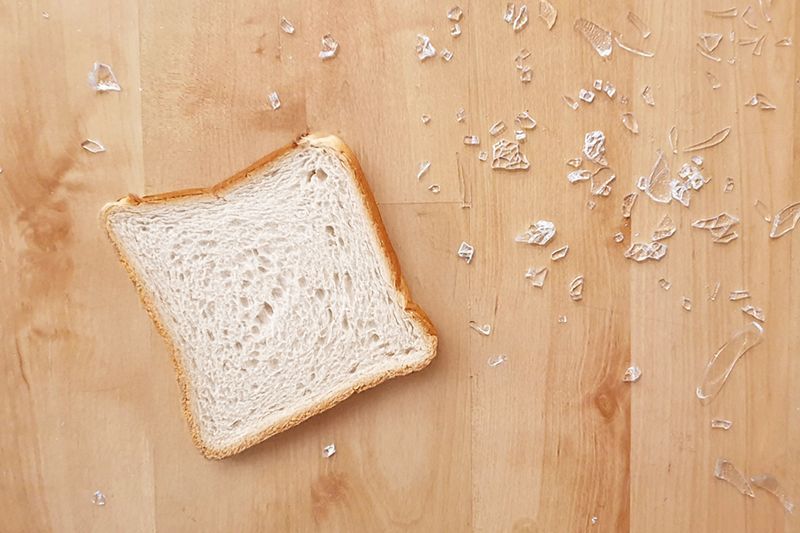 A piece of sliced white bread on a wood floor next to shards of broken glass.