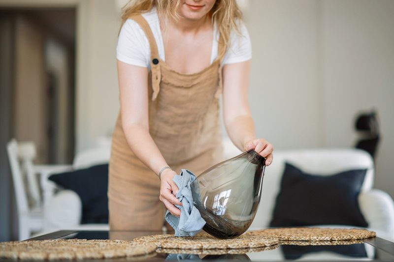 Blonde woman in tan overalls cleaning a large, gray glass vase with a soft cloth.