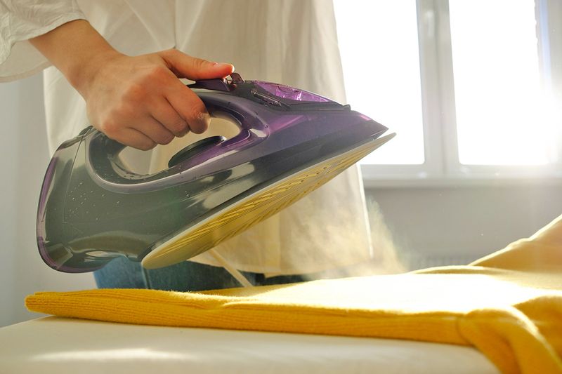 Anonymous person holding a purple iron over a yellow sweater on an ironing board.