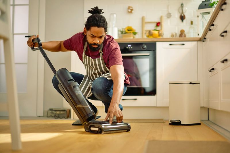 Bearded man inspecting his vacuum cleaner while crouched down in the kitchen.