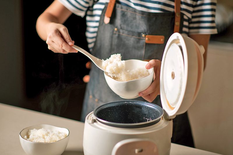 Anonymous person wearing an apron scooping white rice out of a rice cooker and into two bowls.