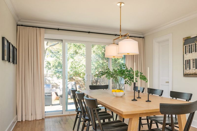 A light-colored dining room with an oak table, black chairs, and pleated curtains that go across sliding doors.