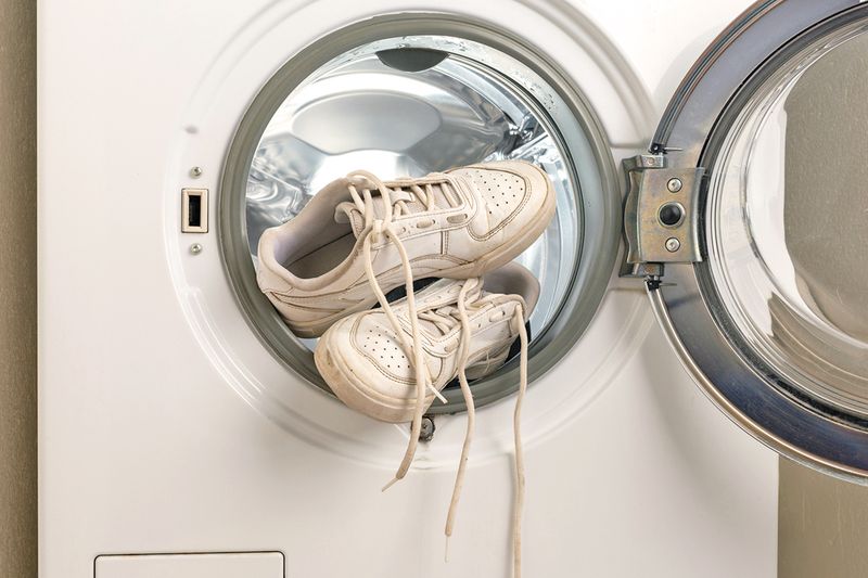 A dirty pair of white sneakers perched on the opening of a washing machine with the door open.