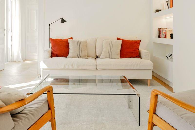 A white room with a loveseat, a glass coffee table, two chairs, a bookshelf, and pops of red.
