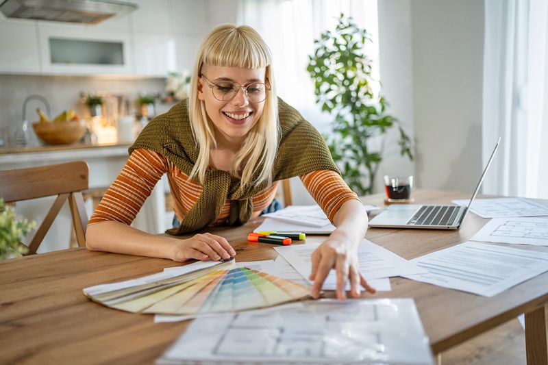 Young woman with blonde hair and micro bangs looking at paint swatches and a floorplan on a wooden kitchen table. Next to her is an open laptop.
