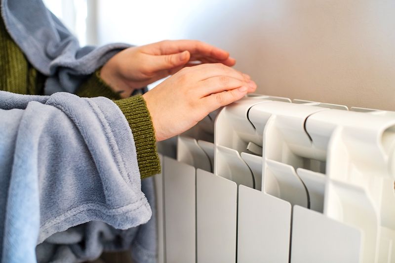 Anonymous person wrapped in a blanket putting their hands over a radiator to warm them.