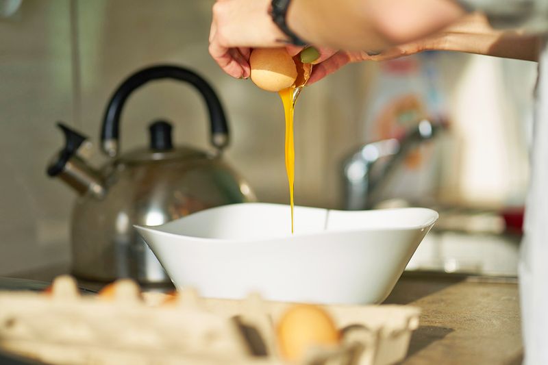 Anonymous person cracking an egg into a bowl. Next to the bowl is an open carton with a few remaining eggs.