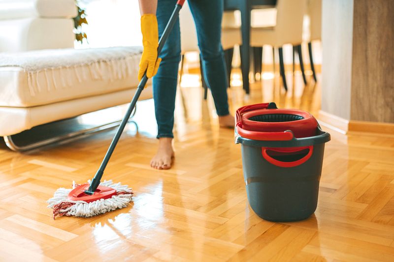 An anonymous, barefoot woman wearing skinny jeans and yellow cleaning gloves. She is mopping hardwood floors in a living room.