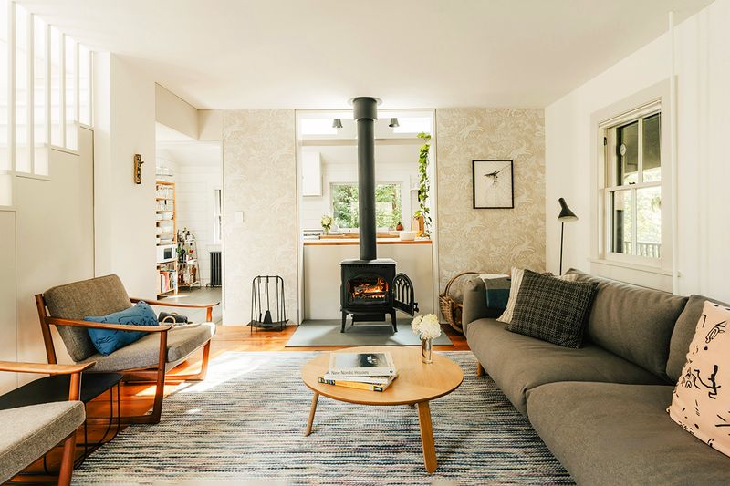 A cozy-looking living room with a wood stove, patterned wallpaper, comfy gray furniture, and a woven rug.