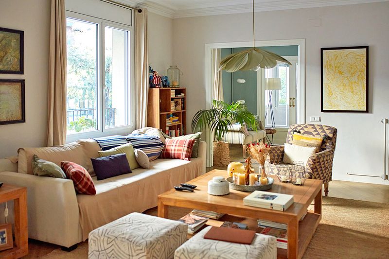 A living room furniture arrangement with a sofa, armchair, coffee table, two poufs, a side table, and a chandelier. The sofa has a slipcover and is covered in throw pillows.