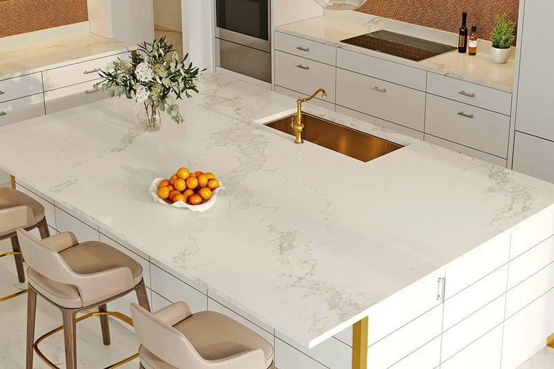 A pristine white marble-topped kitchen island with a gold sink and faucet and three barstools pulled up to it.
