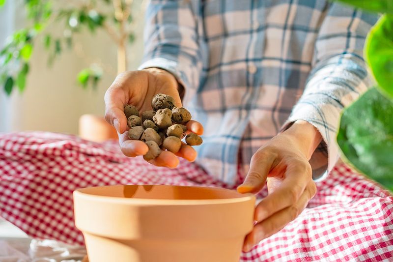 Anonymous person wearing a plaid shirt adding a handful of rocks to a terra cotta plant pot.