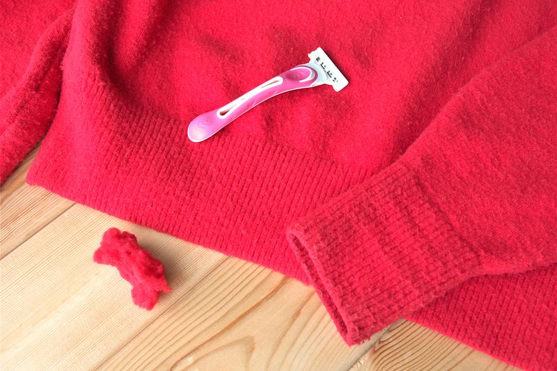 A pink disposable razor on top of a red, pilled sweater. Next to the sweater is a pile of red fuzz.
