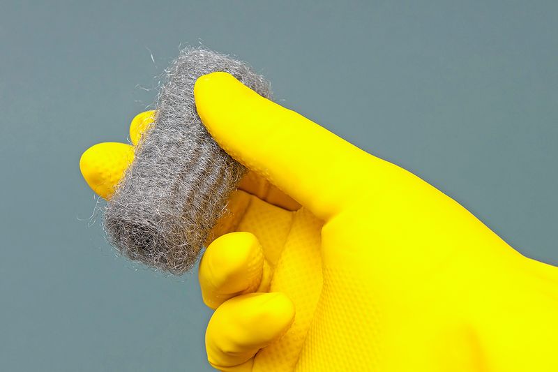 Anonymous person wearing a yellow cleaning glove and holding steel wool in between their fingers.