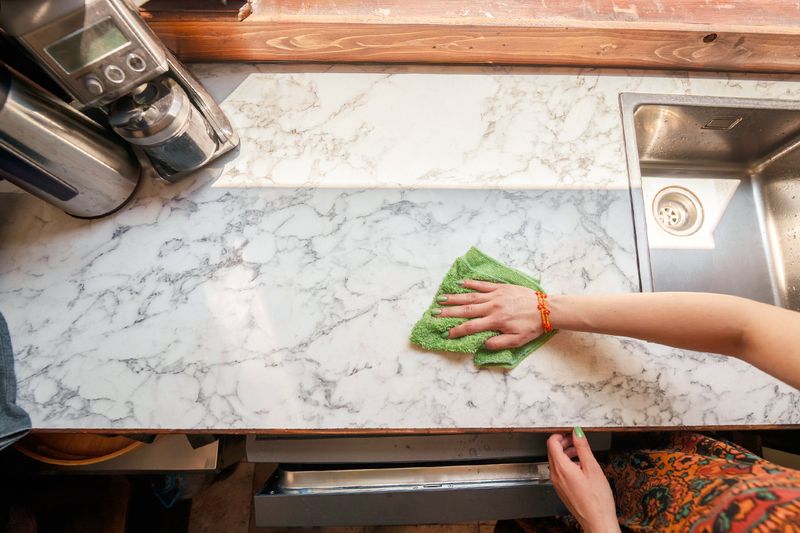 Anonymous person wiping down a marble countertop with a microfiber cloth. On the counter is a coffee maker and a kettle, both of which are stainless steel.