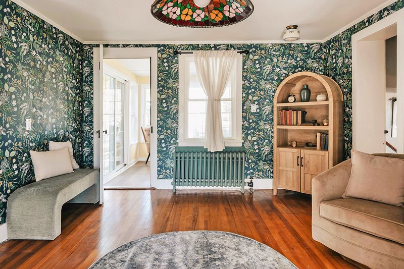 A room with teal botanical wallpaper, a round patterned rug, a Tiffany-style lamp shade, and a few pieces of furniture.