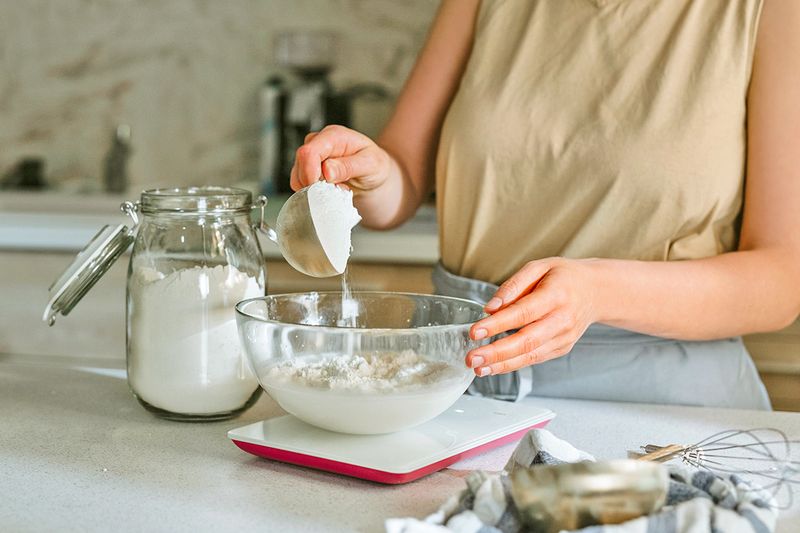 Anonymous woman measuring flour into a glass mixing bowl. The bowl is on a scale.