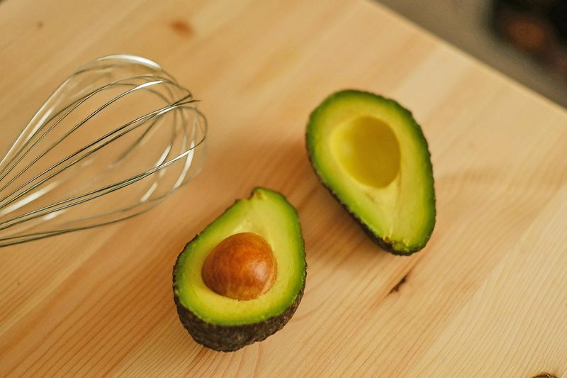 Both halves of a ripe, cut avocado on a wooden cutting board next to a wire whisk.