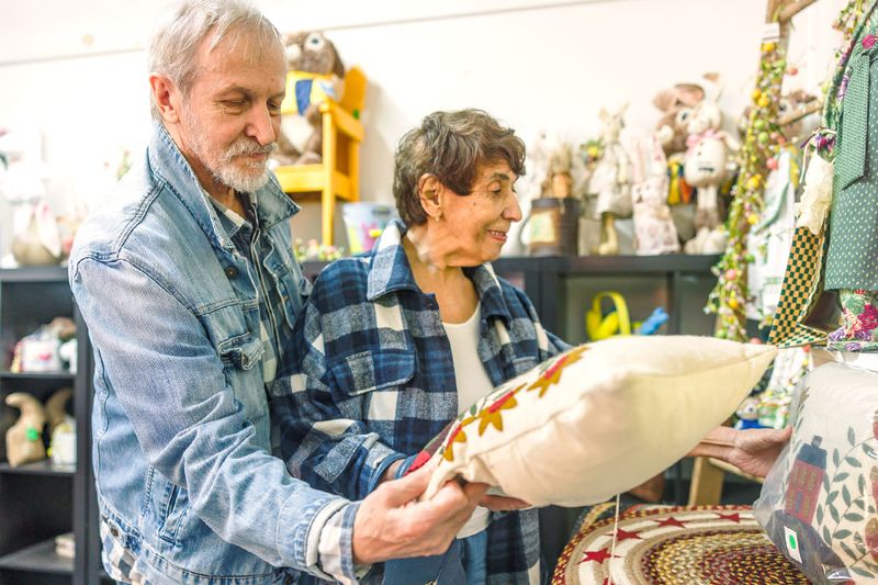 An older couple browsing a thrift store. The man, wearing a jean jacket, is looking at a patterned throw pillow.