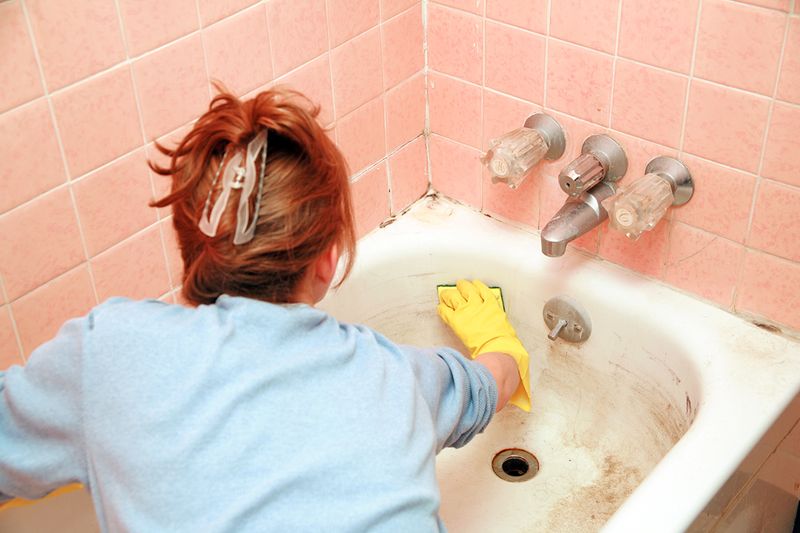 Anonymous woman cleaning a grimy bathtub. She is wearing yellow cleaning gloves and holding a sponge.