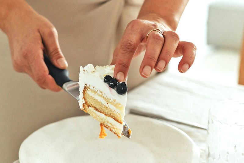 Anonymous person putting a slice of cake on a plate using a knife to serve it. The cake is yellow with white icing and blueberries on top.