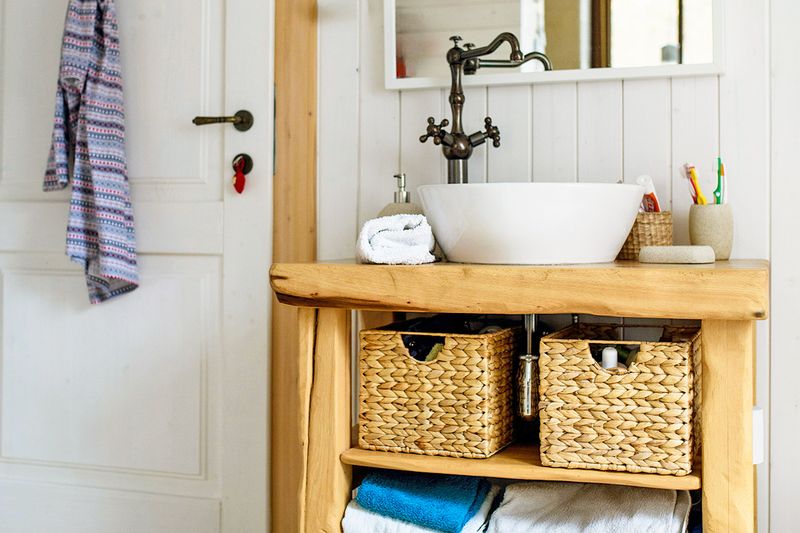 A bathroom with a natural wood vanity and a vessel sink. On the counter are toothbrushes, toothpaste, soap, and a towel. Underneath, there are woven storage baskets and folded towels.