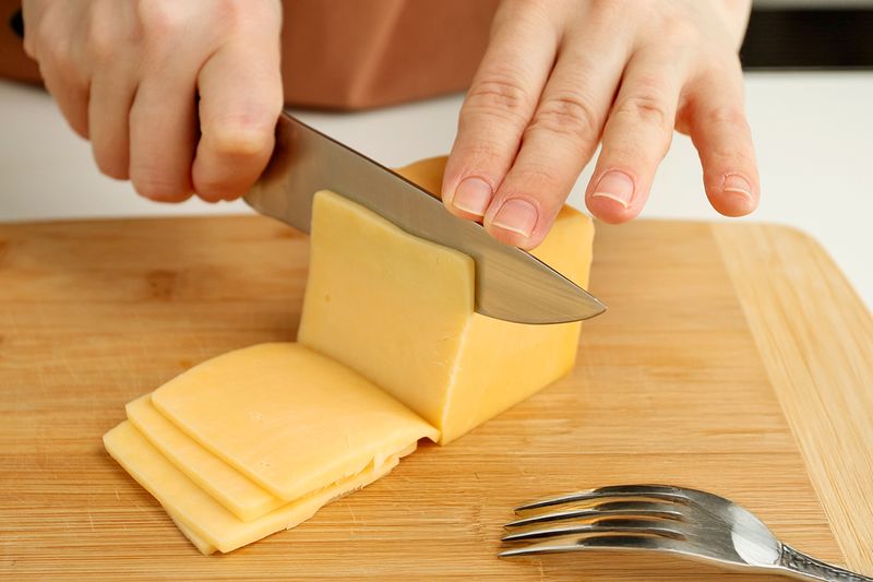 Anonymous person slicing cheddar cheese on a wooden cutting board.