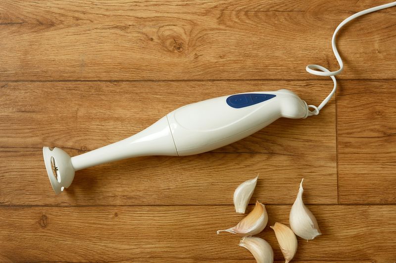 An immersion blender laying flat on a wooden countertop next to a few cloves of garlic.