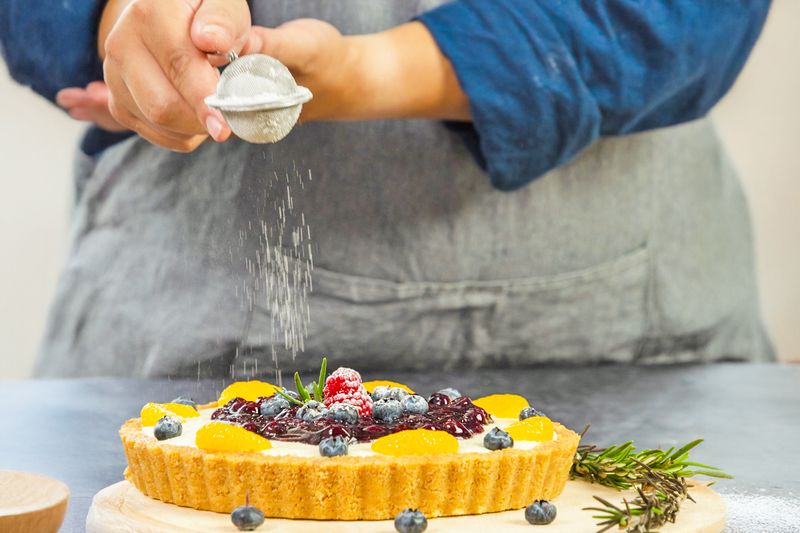 An anonymous person using a snap-ball tea infuser to sprinkle powdered sugar over a fruit tart.