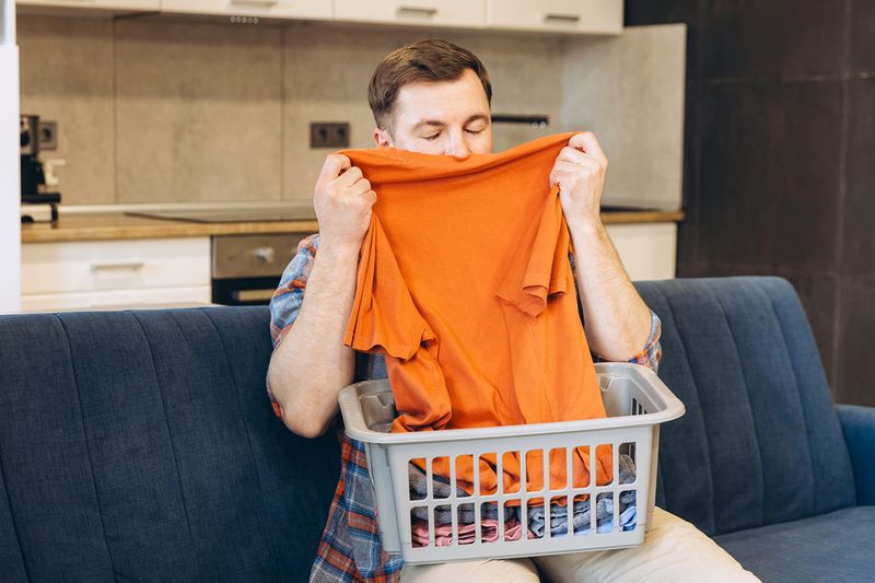 A young man smelling an orange T-shirt from a laundry hamper that's sitting on his lap.