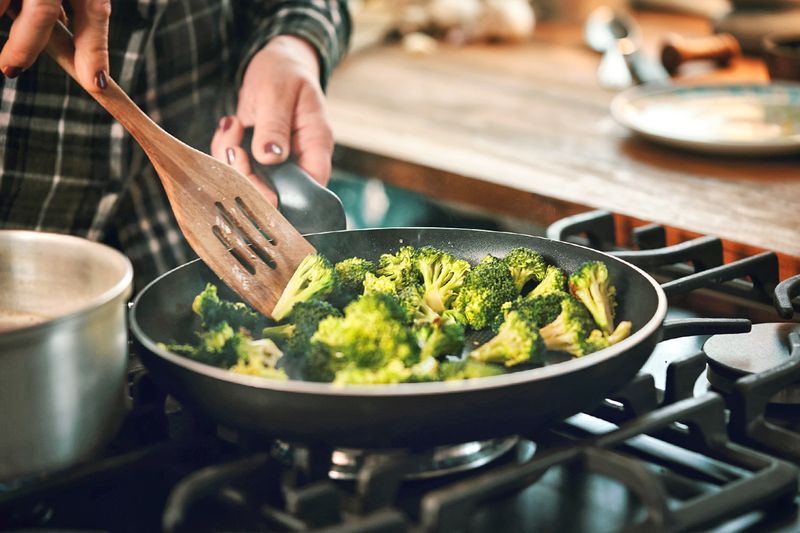 Anonymous person stir-frying broccoli in a skillet using a wooden spatula.