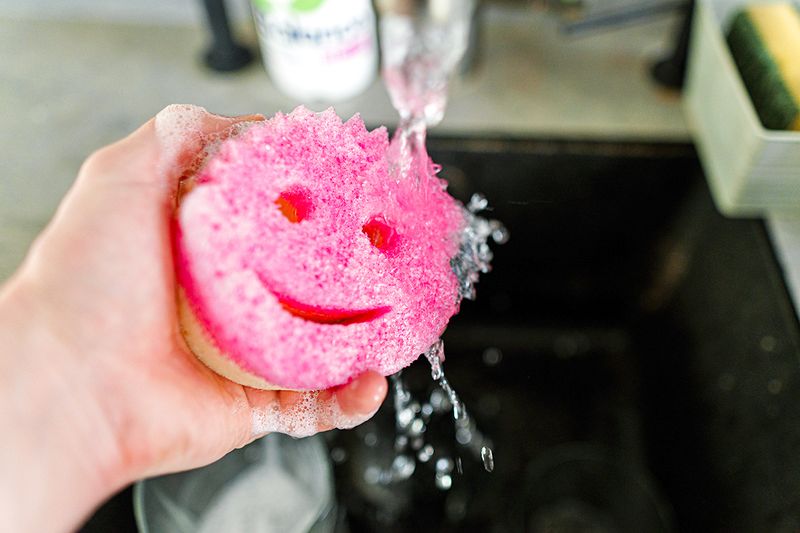 A pink Scrub Mommy sponge under a running faucet.