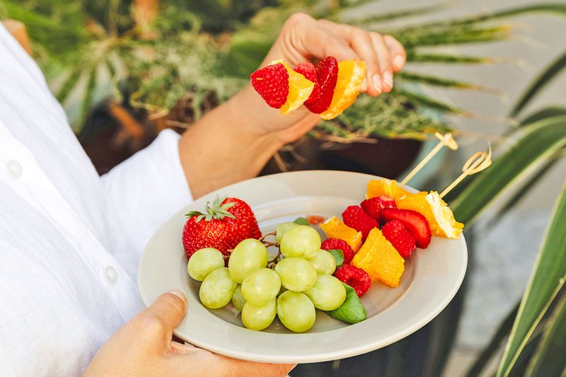 Anonymous person holding a plate full of green grapes and skewers of oranges, strawberries, and raspberries.