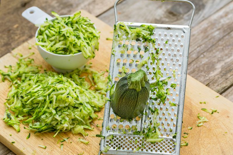 A wooden cutting board, a grater, and a measuring cup, all full of grated zucchini.