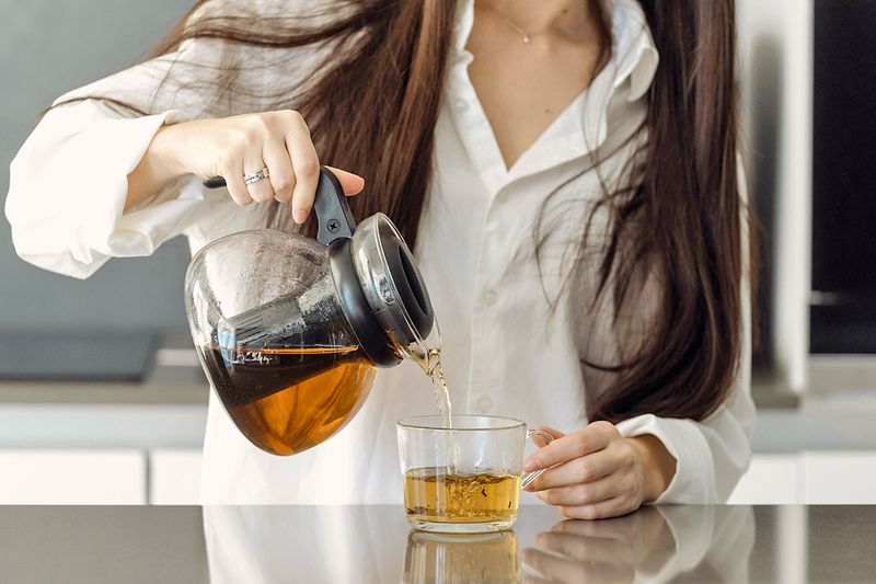 Anonymous woman pouring tea out of a glass kettle into a glass mug.