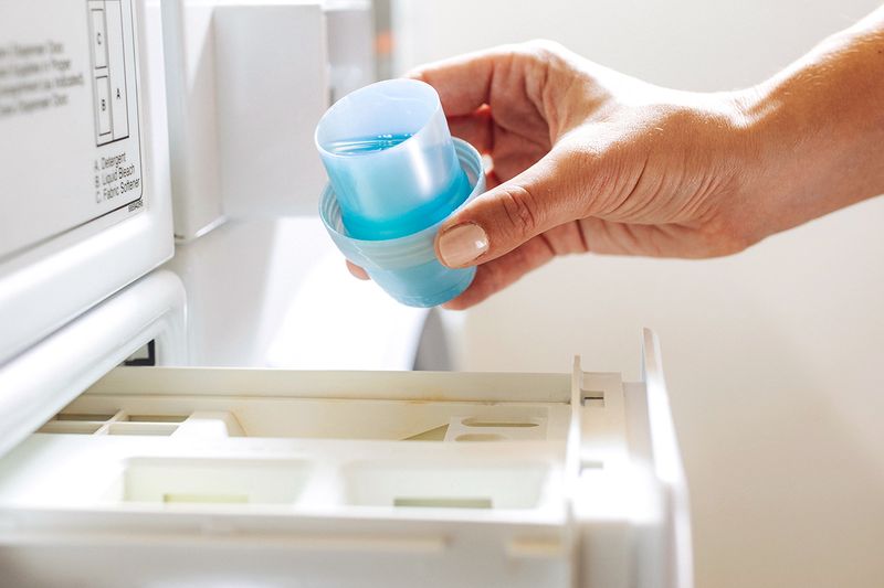 Anonymous person pouring a cap of blue laundry detergent into the washing machine's detergent drawer.