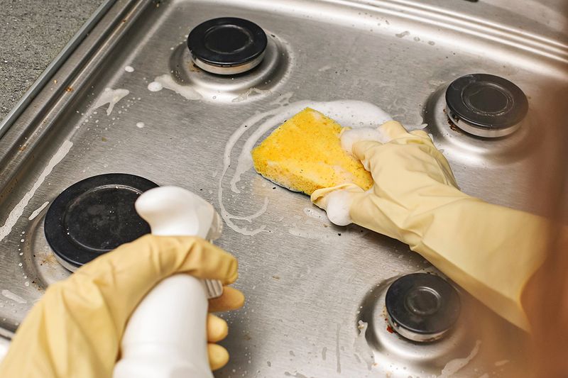 Anonymous person cleaning a metal stovetop with cleaner and a sponge.