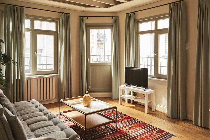 An apartment living room with opened blackout curtains hanging from every window.
