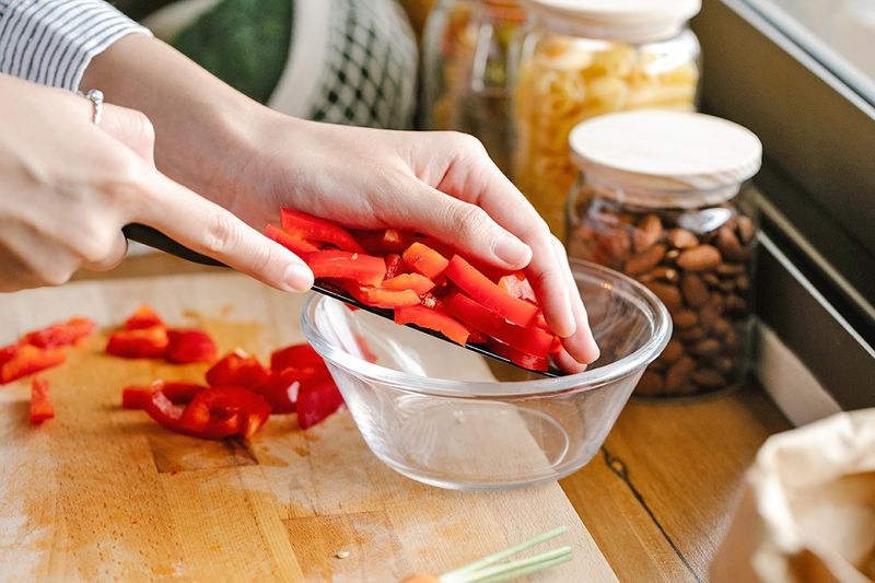 Anonymous person transferring slices of red bell pepper from their knife to a clear glass bowl.