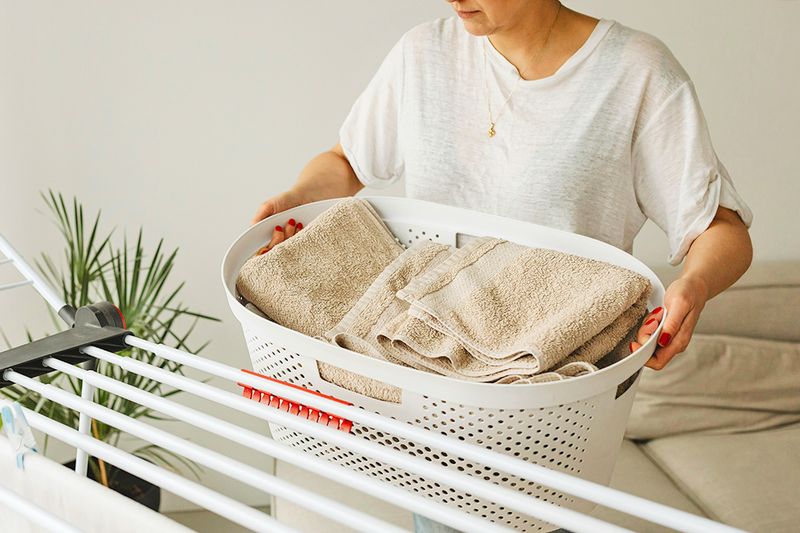 Anonymous woman holding a laundry basket full of folded towels in front of a drying rack.